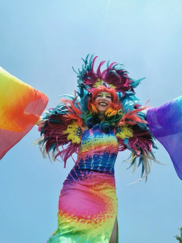 Joyful Pride stilt-walker from below with outstretched rainbow silk arms and feathered headdress against blue sky