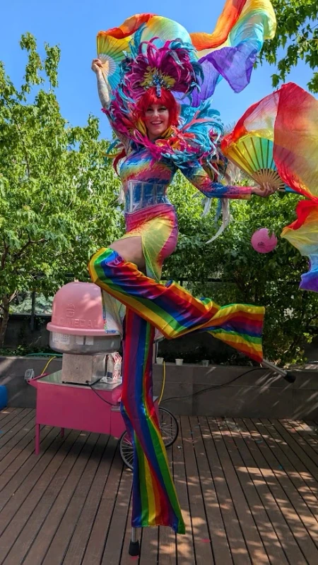 Full-length Pride stilt-walker in rainbow striped pants with flowing silk fans and feathered headdress at rooftop event