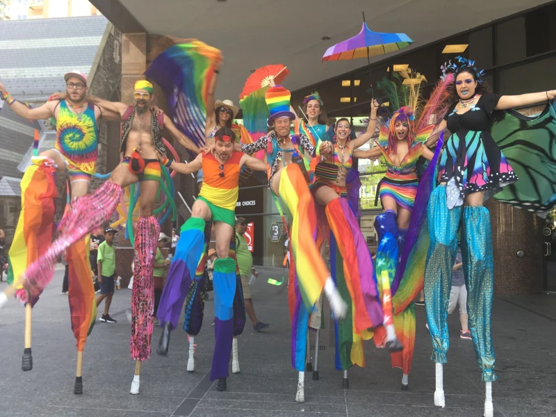 Wide shot of Pride parade with multiple rainbow stilt performers towering over enthusiastic crowds