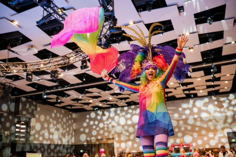 Rainbow Pride stilt performer with purple feathers and silk fan veils at indoor corporate event with spotlight