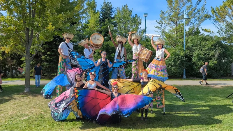 Full TallBeat troupe group photo with butterfly wing ground performers at park, stilt drummers towering above with branded drum