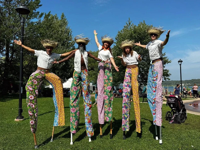 Five TallBeat stilt-walkers posing with open arms by the waterfront at a summer lakeside festival