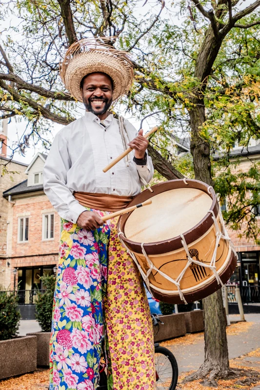 TallBeat drummer on stilts playing alfaia drum with joyful expression, wearing straw hat and colourful floral pants