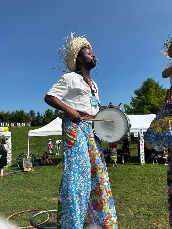 TallBeat drummer performing solo on stilts with snare drum at summer festival, wearing blue floral pants and straw hat