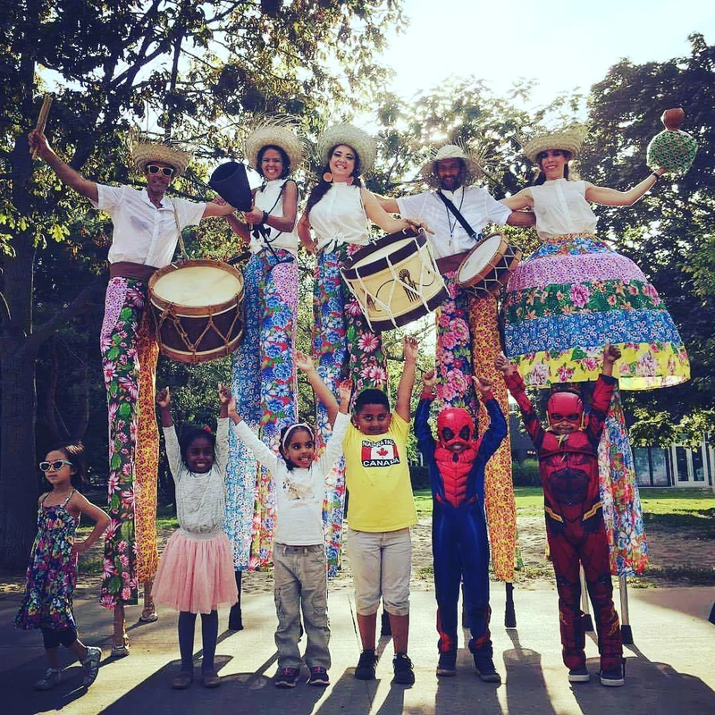 TallBeat troupe performing for excited children at park festival, kids dancing below the towering stilt drummers